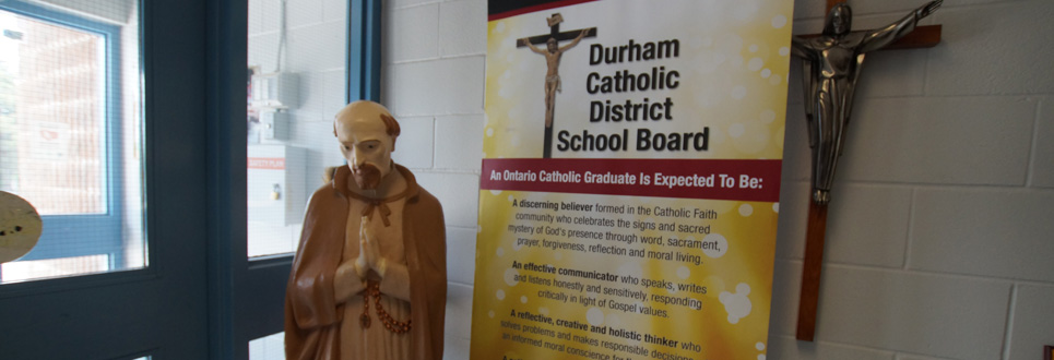 Stand up sign stating the Catholic Graduate Expectations beside a statue of St. Isaac Jogues.